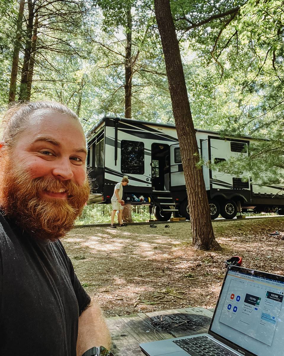 Andy working on a laptop at a campsite picnic table with the family fifth wheel behind him